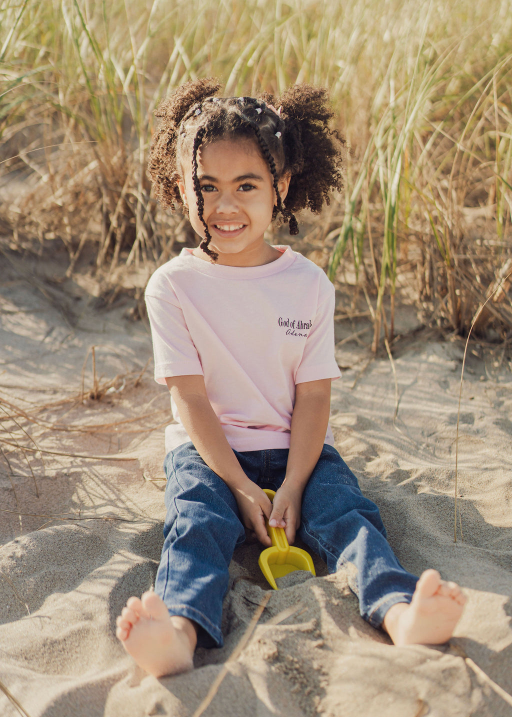 Child sitting on sand with grasses in the background, wearing a pink Jeremiah 1:5 t-shirt and blue jeans.