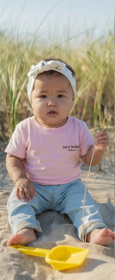 Baby sitting on sand wearing a pink Jeremiah 1:5 shirt and white headband. 
