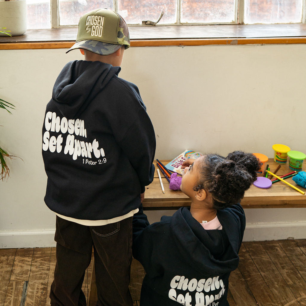 Two children wearing matching black hoodies with 'Chosen Set Apart.' printed in white on the back, standing and sitting in a room with a wooden floor and large window.