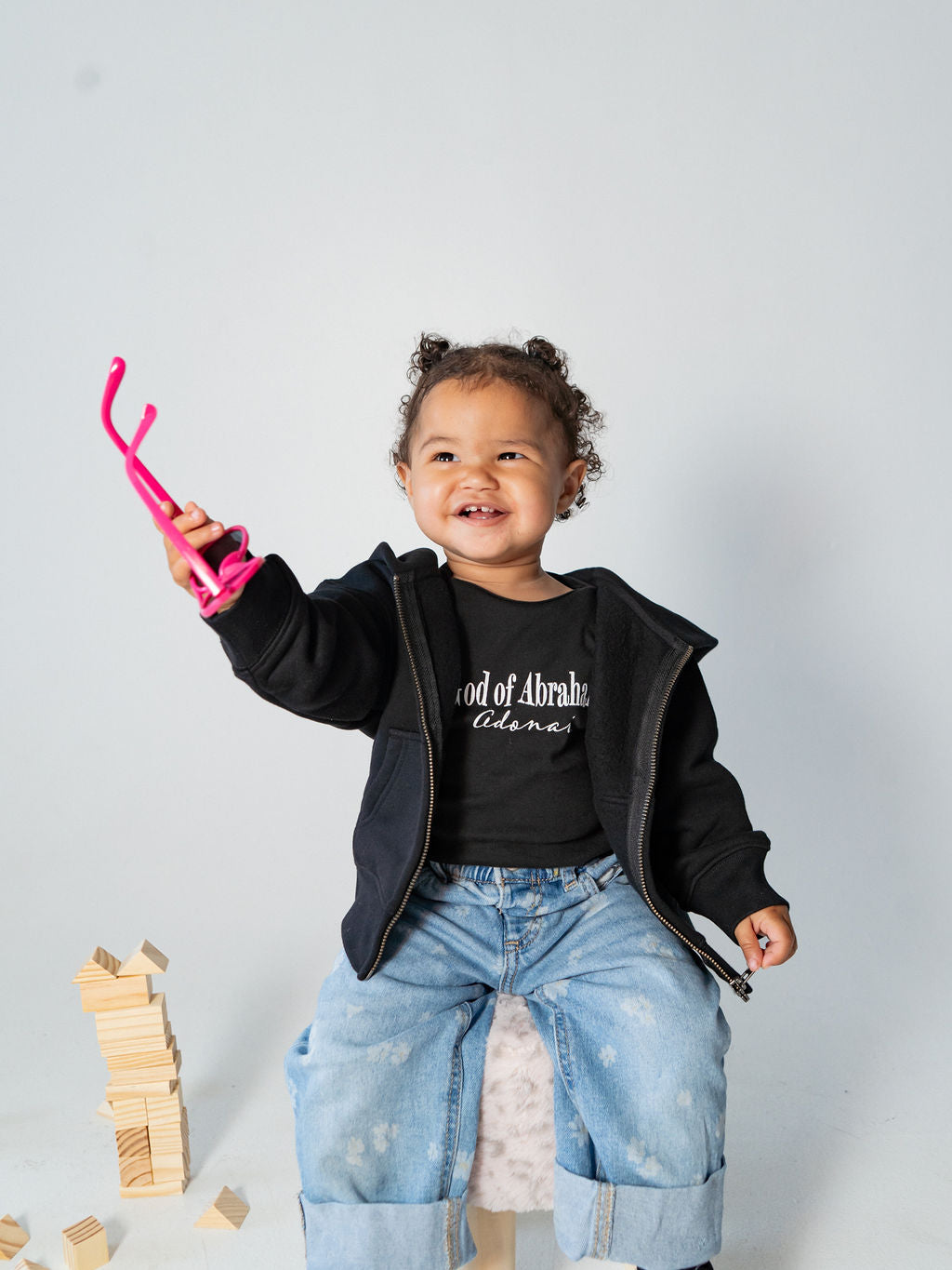 A smiling toddler girl with curly hair stands on a stool wearing a black Lion of Judah zip-up hoodie over a black graphic t-shirt and light blue jeans. She holds a pair of bright pink safety glasses in one hand, with wooden blocks scattered on the floor against a light gray studio background.