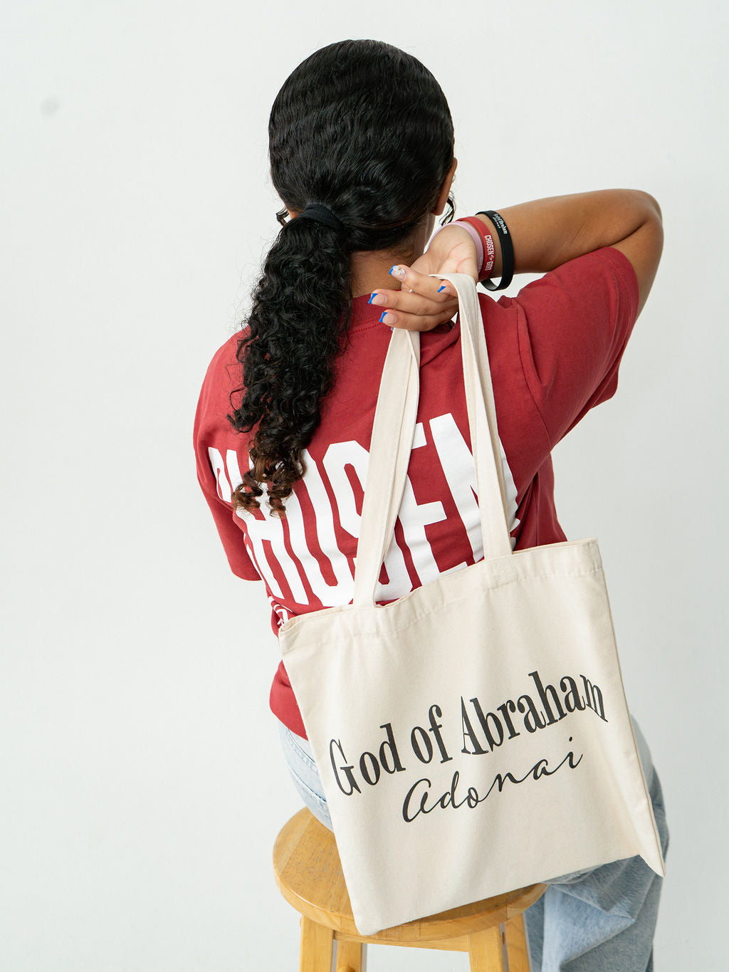 A person viewed from behind wearing a red t-shirt and light blue jeans, holding a cream canvas tote bag over their shoulder. The tote bag has black text that reads 'God of Abraham Adonai'.