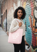 Woman holding a pink tote bag with text in front of a graffiti wall.